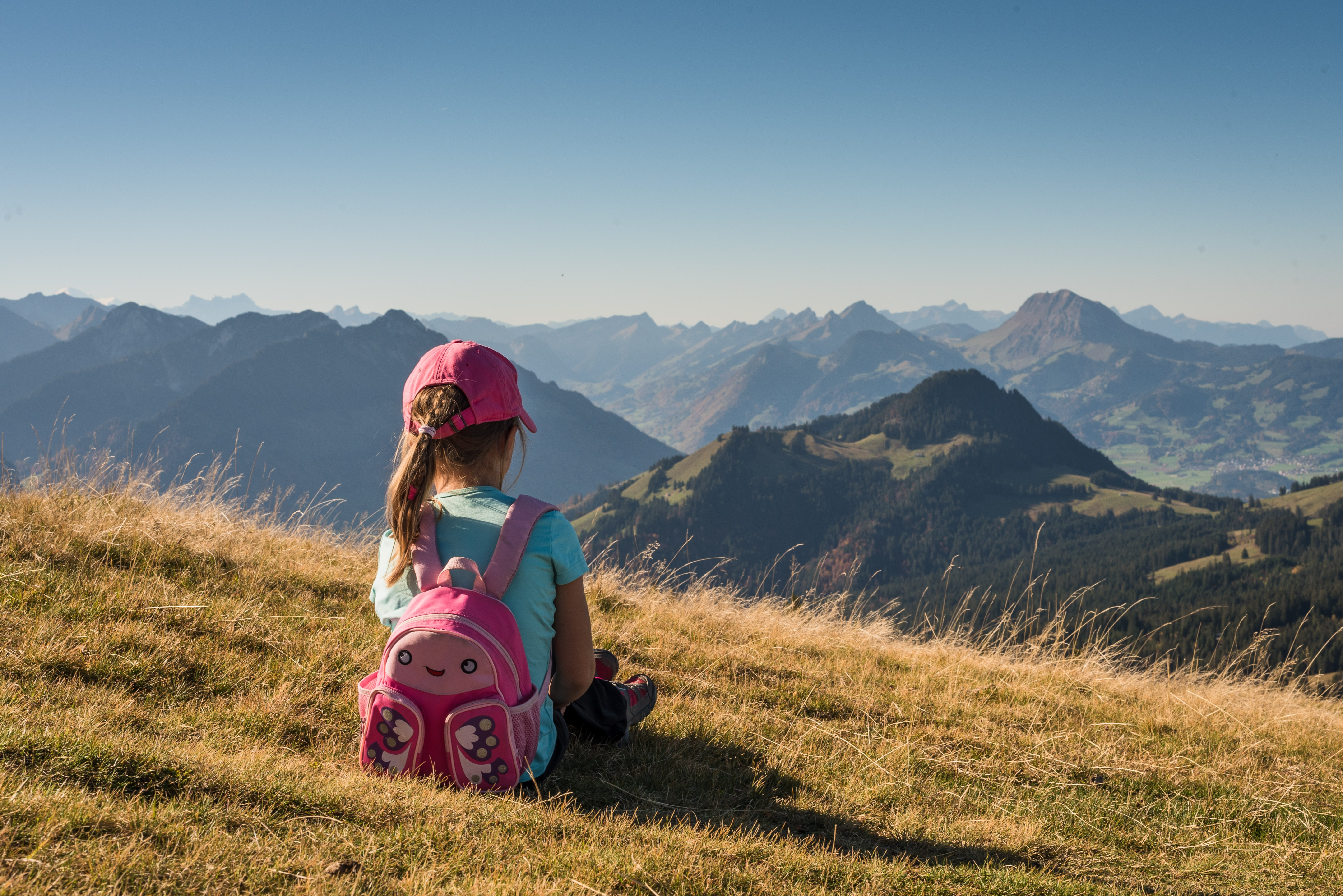 A young girl sits on a hill with a pink hat and backpack